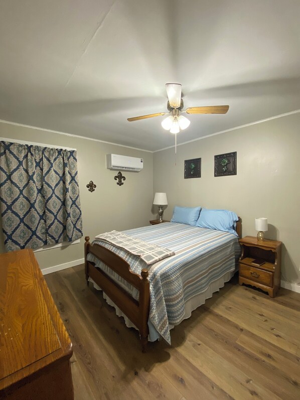 Un dormitorio con cama de madera, ventilador de techo, mesita de noche con lámpara y ventana con cortinas estampadas.