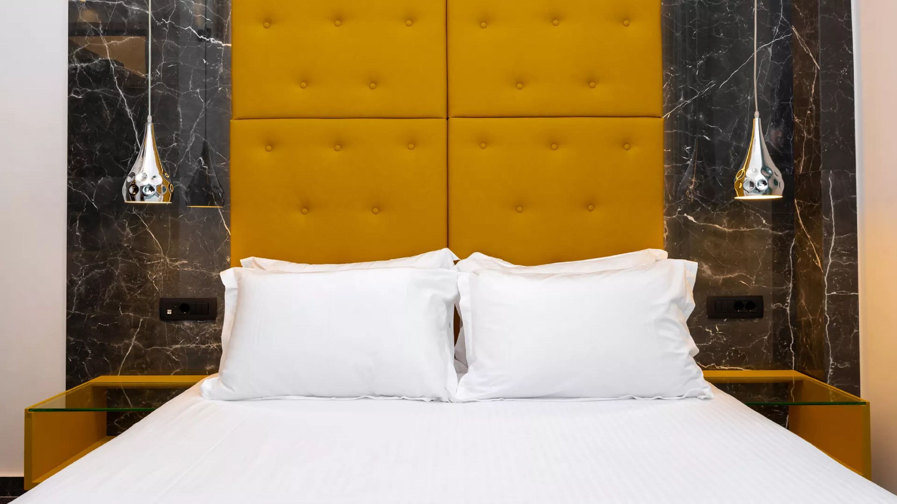 Un lit avec du linge de lit blanc, une tête de lit jaune à motifs capitonnés et deux tables de chevet avec des plateaux en verre.