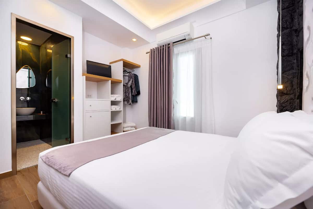 A modern hotel room with a large bed, a walk-in closet, a bathroom with a mirror and sink, and a stone accent wall.