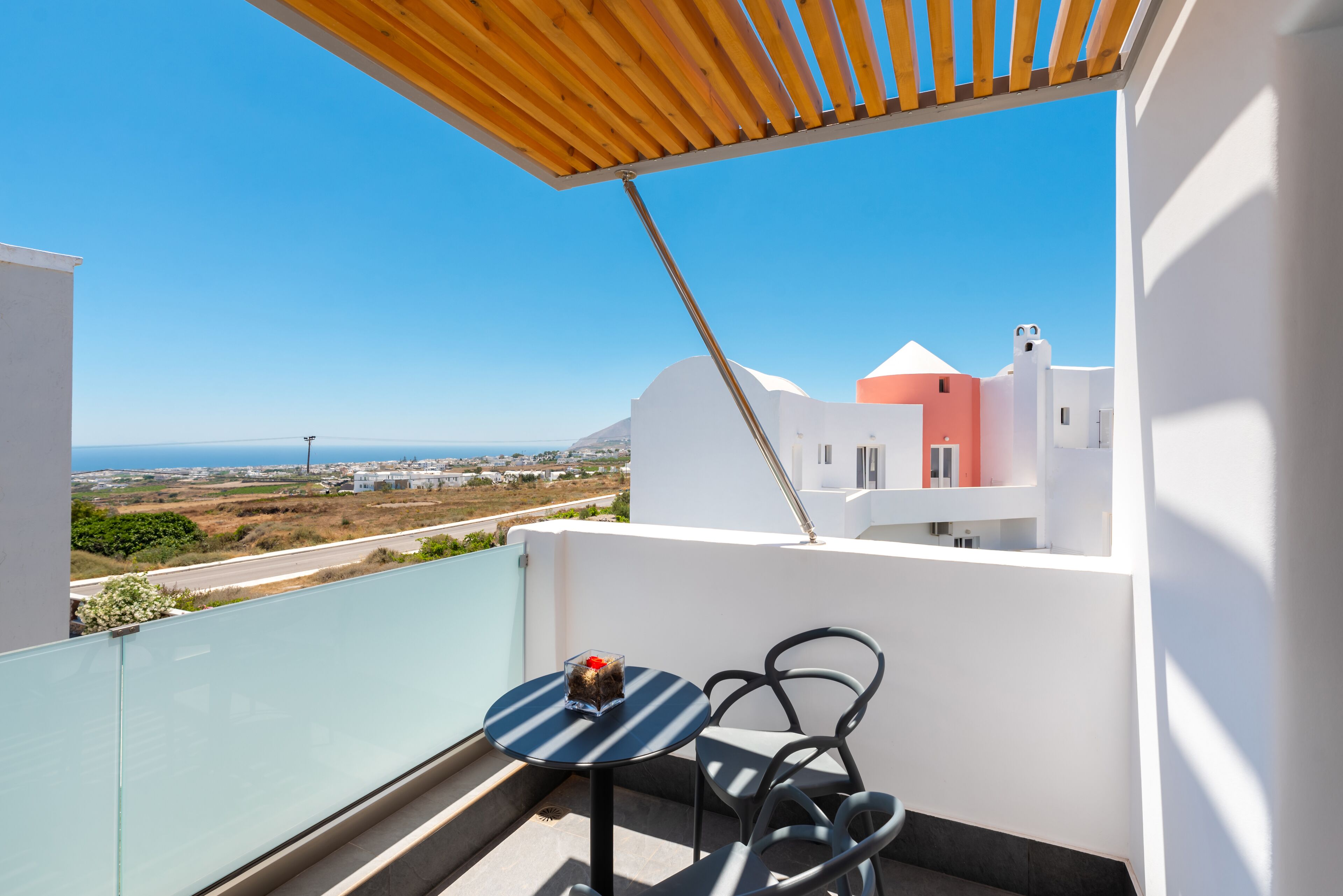 A balcony with a view of the sea and buildings, a table with chairs, and a wooden pergola.
