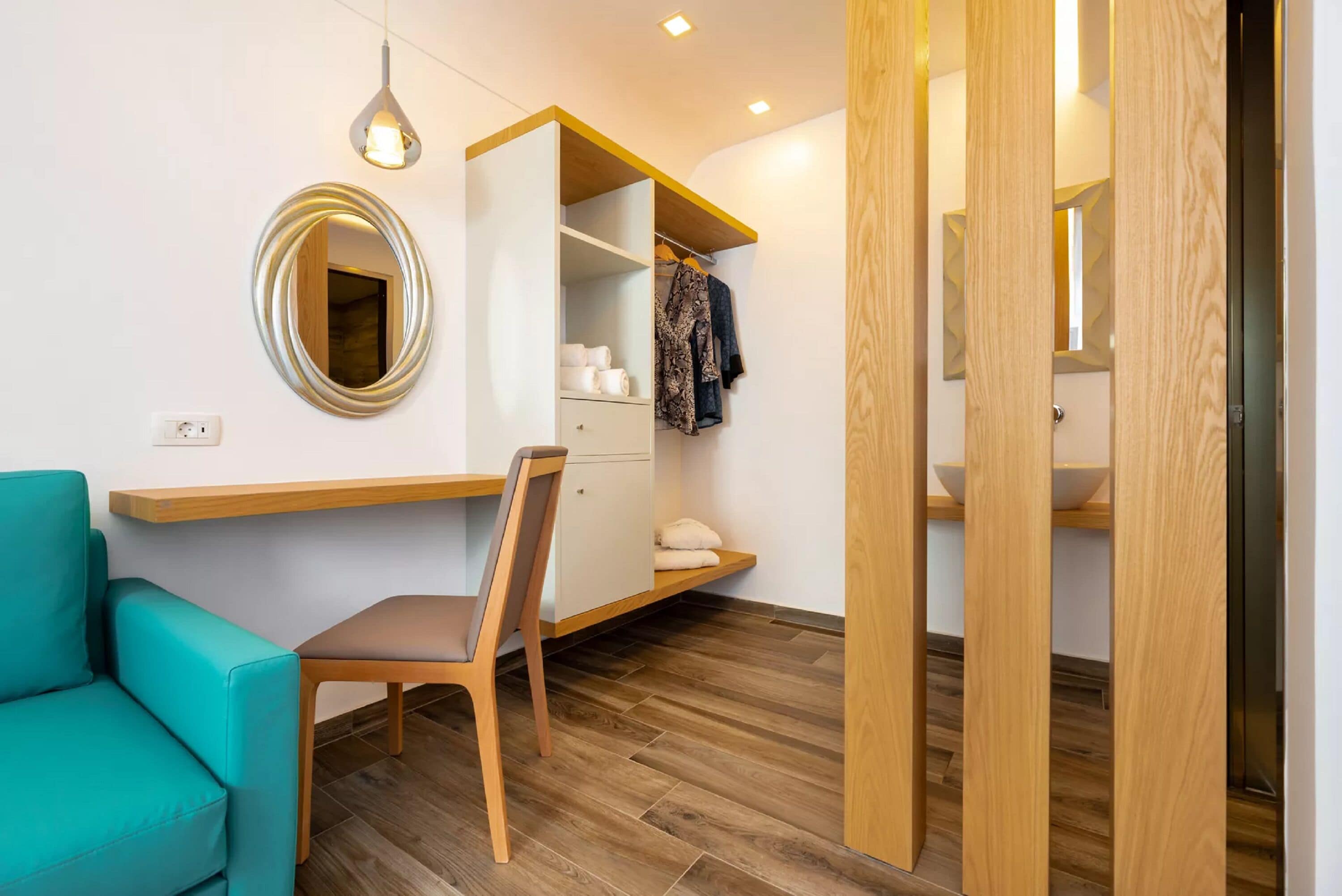 A modern room with a teal sofa, wooden desk, and a wardrobe with hanging clothes.