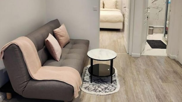 Una sala moderna con un sofá gris, una mesa redonda y una alfombra pequeña. Se ve un dormitorio con una cama y un cuadro a través de una puerta abierta.
