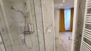 Un baño moderno con cabina de ducha de vidrio, paredes de mármol y un pasillo visible que conduce a una habitación con cortinas amarillas.