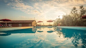 Una piscina con agua cristalina que refleja el cielo y los árboles, rodeada de sillas de descanso y sombrillas.