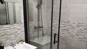 A modern bathroom with a glass-enclosed shower, a white sink with a mirror, and a tiled floor. - Modern, fully-equipped apartment (L'Isle-Jourdain)