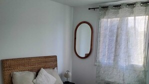 A bedroom with a single bed, a bedside table, a lamp, a mirror, and a window with a patterned curtain. - Modern, fully-equipped apartment (L'Isle-Jourdain)