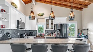 A modern kitchen with white cabinets, a large island with bar stools, stainless steel appliances, and hanging pendant lights. - Beachfront 3B/3B Fishing Oasis (Ramrod Key)