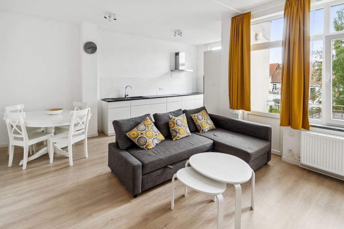 Una sala moderna con un sofá gris, sillas blancas, una mesita pequeña y una cocina con armarios blancos y encimera negra.
