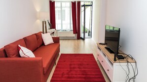 Un salon moderne avec un canapé rouge, un téléviseur posé sur un meuble en bois et une vue sur l’extérieur grâce à une porte vitrée.