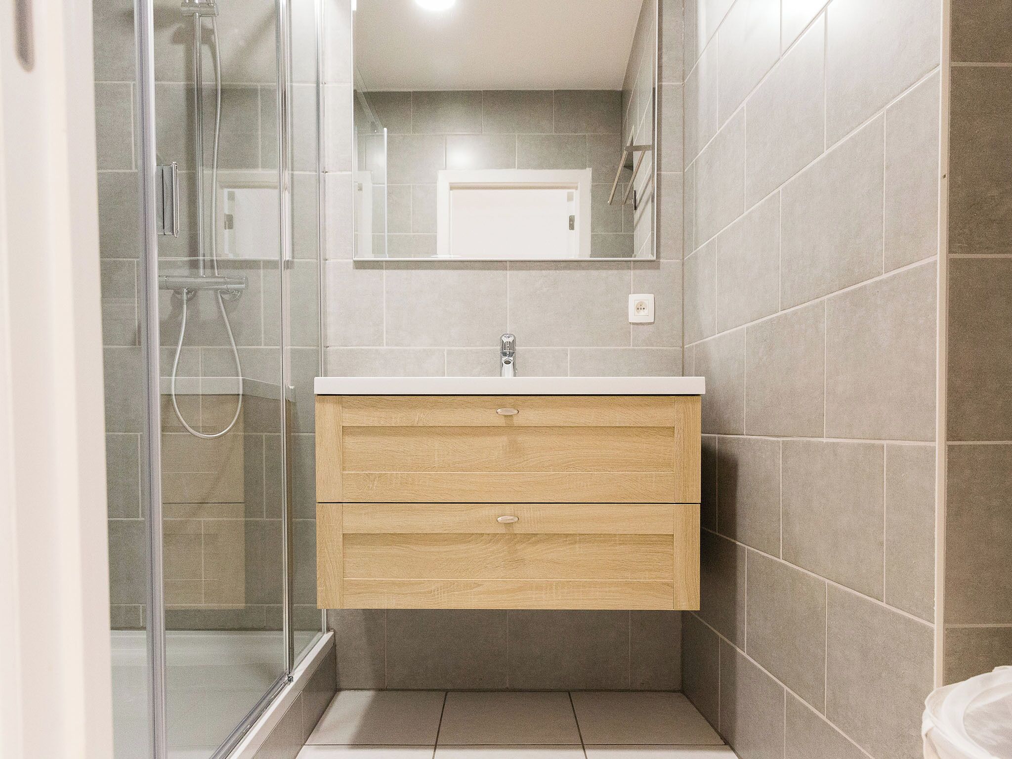 Een moderne badkamer met een douchecabine van glas, een houten wastafelmeubel met een wastafel en een spiegel erboven.