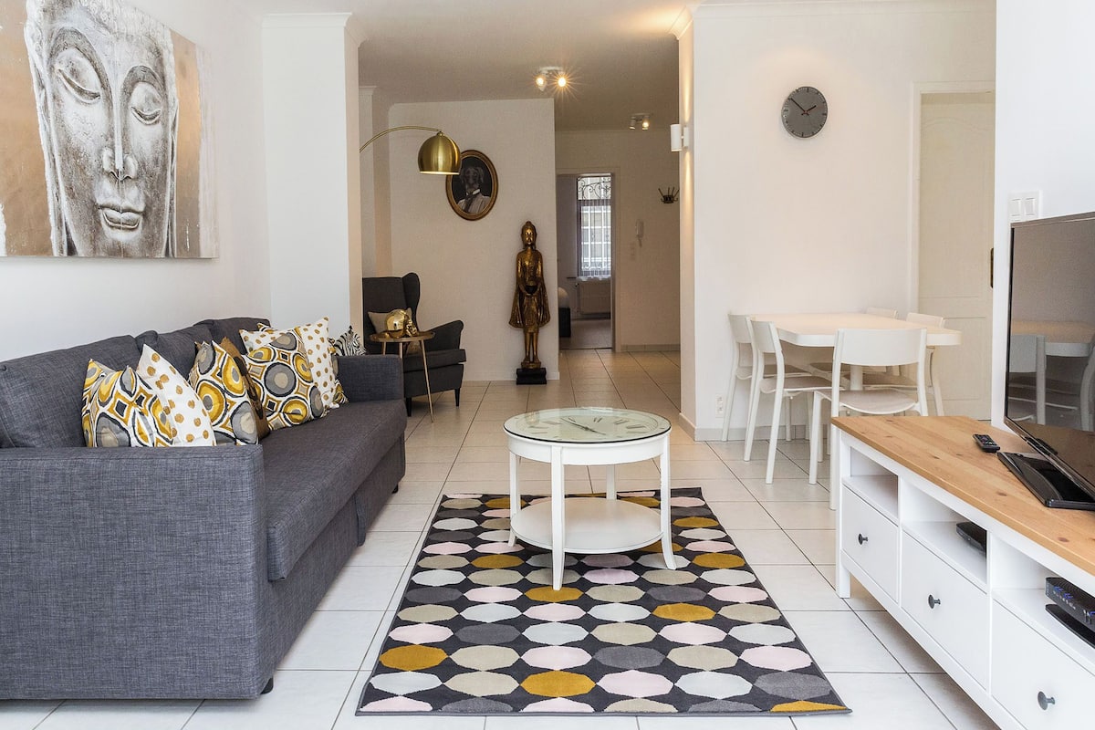 Una sala moderna con un sofá gris, una mesita de centro y un tapiz estampado. Se observa un comedor con una mesa y sillas blancas, y una zona de cocina con un refrigerador y un televisor.