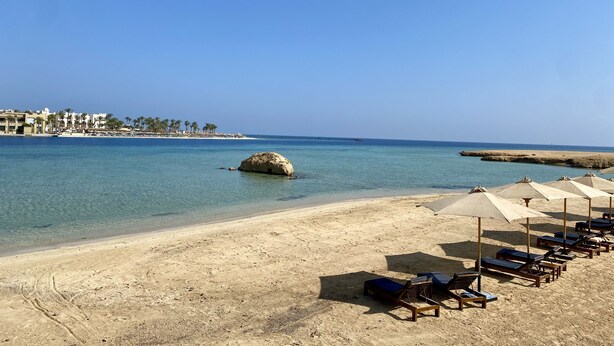 Une plage à l’eau d’un bleu cristallin, un promontoire rocheux et des rangées de transats avec des parasols.