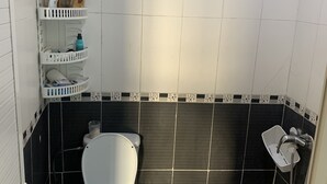 Une petite salle de bain avec des toilettes, une douche et un lavabo. On y trouve des étagères avec des articles de toilette et une serviette accrochée au mur.