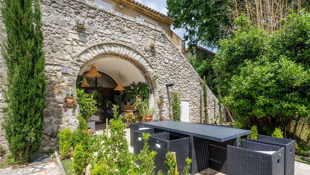 Un bâtiment en pierre doté d'une grande porte en arc de cercle, d'un espace extérieur aménagé avec des meubles en osier et d'une terrasse carrelée.