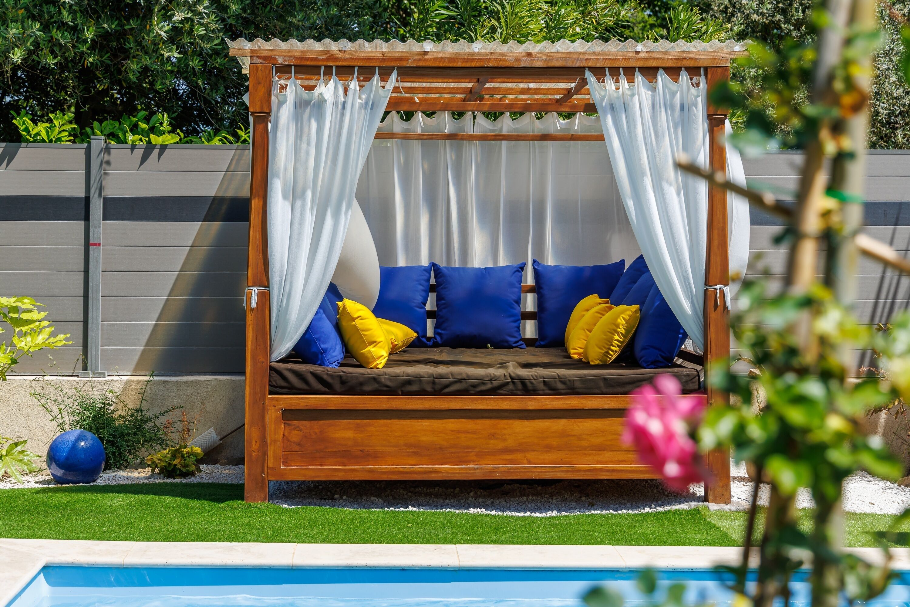 A wooden outdoor bed with blue and yellow cushions, white curtains, and a pool in the background.