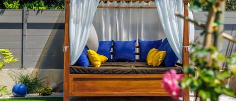 A wooden outdoor bed with blue and yellow cushions, white curtains, and a pool in the background.