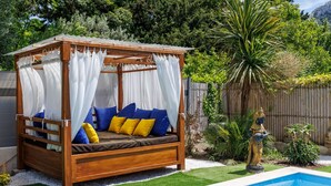 Un lit d’extérieur en bois, agrémenté de rideaux blancs, de coussins bleus et jaunes, avec une piscine en arrière-plan.