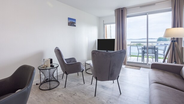 Una habitación de hotel moderna con un amplio ventanal, un televisor de pantalla plana, un sofá gris y una mesita auxiliar.
