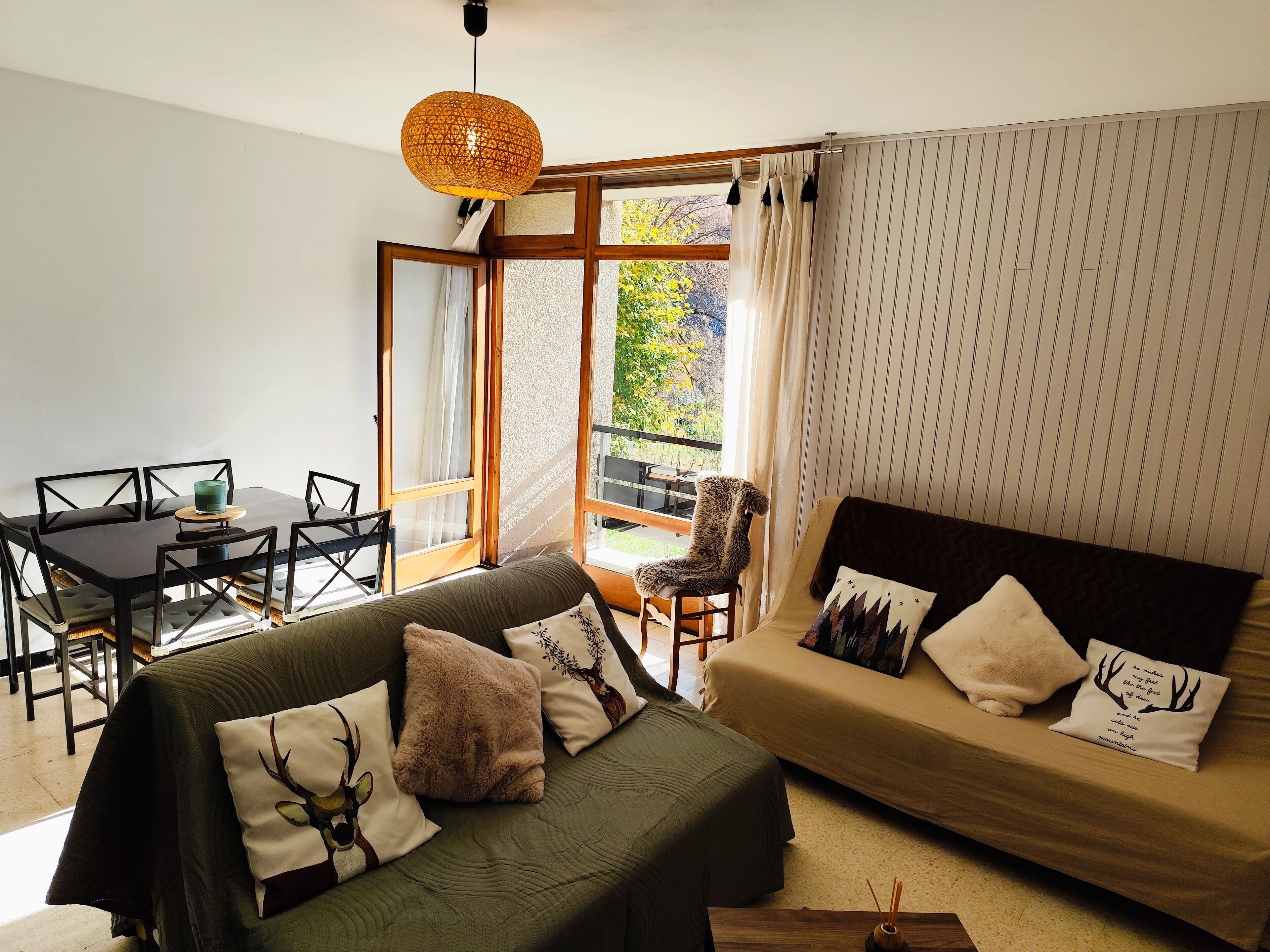Un salon chaleureux avec un canapé, une table à manger et une chaise. Il y a une fenêtre avec des rideaux, un luminaire suspendu et une vue sur la verdure à l’extérieur.