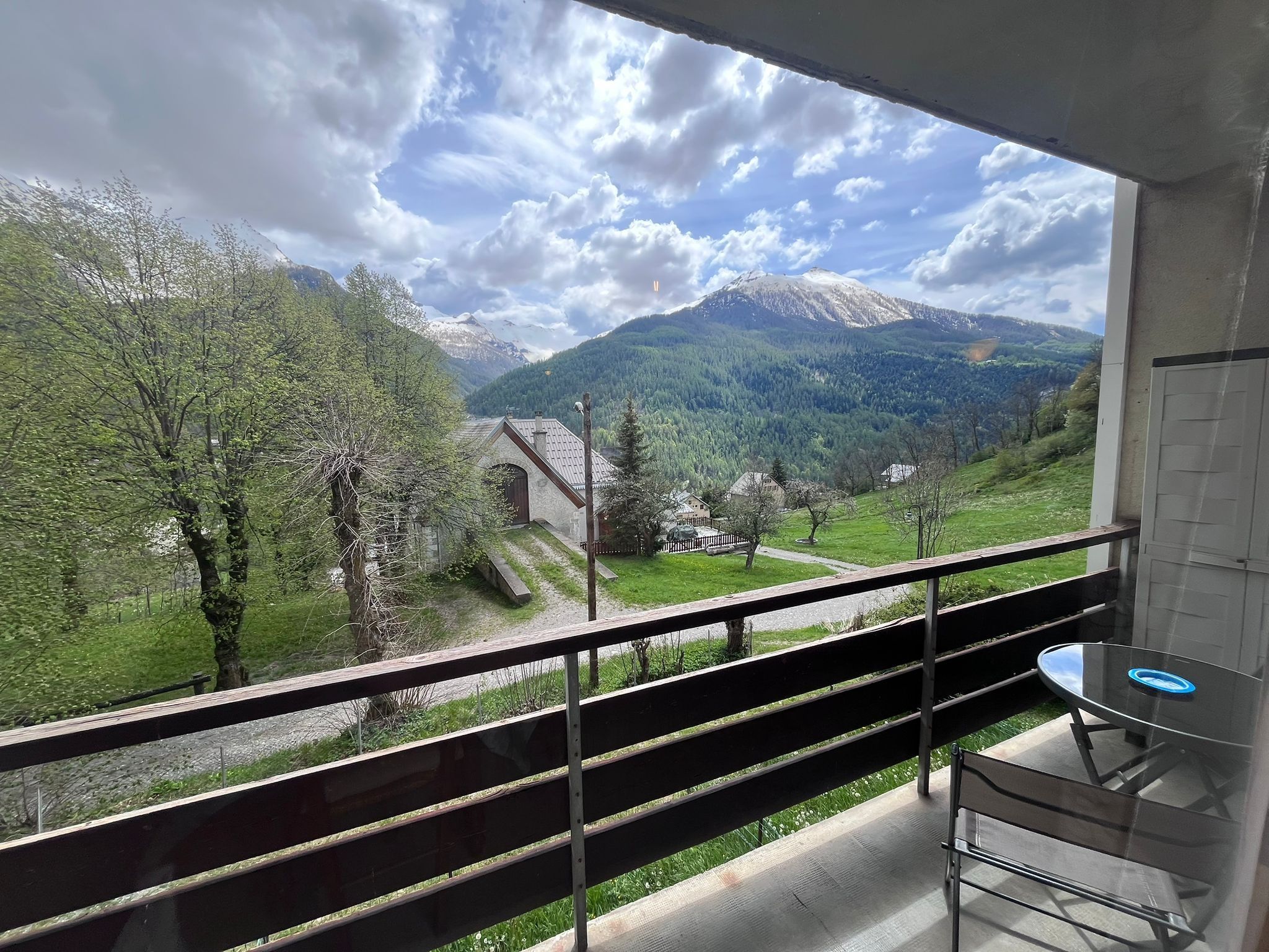 Un balcone con tavolo e panca in vetro, affacciato su un paesaggio montuoso con alberi e cielo sereno.