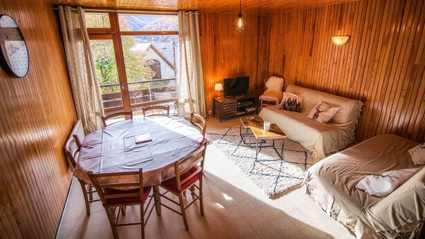 Ein gemütliches Blockhaus-Interieur mit Holzbalkendecke, einem Esstisch mit Stühlen, einer Couch, einem Fernseher und Blick ins Freie.