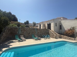 Une piscine entourée d’un mur de pierre et d’un bâtiment doté de fenêtres et de volets, avec des chaises longues et des parasols à proximité.