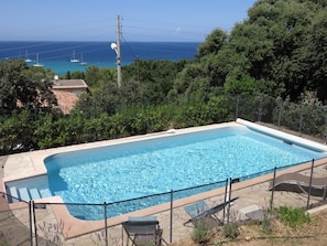 A rectangular swimming pool with clear blue water, surrounded by a stone deck and a metal fence. In the background, there are trees and a view of the ocean with boats.
