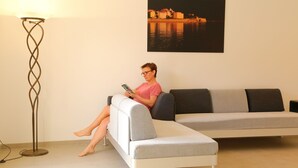 Eine Person sitzt auf einem grauen Sofa, neben einem Stehlampen und einem Bilderrahmen mit einem Blick auf das Wasser an der Wand.