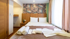 A hotel room with a neatly made bed, a wooden wardrobe, a bedside table with a lamp, and a geometric patterned wall.