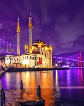 A mosque with two minarets and a large dome, situated by a body of water with a bridge in the background.