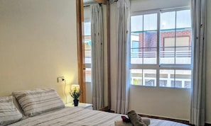A bedroom with a bed, a bedside table with a lamp, a window with curtains, and a view of a building outside.