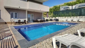 A modern two-story building with a swimming pool, outdoor seating, and a view of a hillside.
