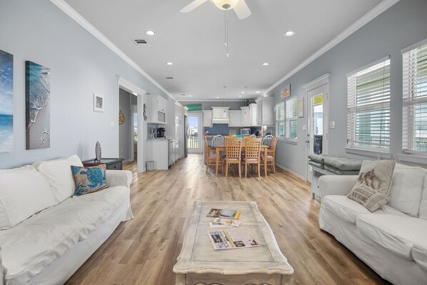 A spacious living room with a white sofa, a coffee table with magazines, and a dining area with wooden chairs and a table.