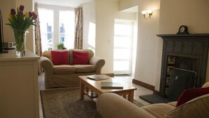 A living room with a sofa, a coffee table, and a fireplace. There is a vase with flowers on a table, and a window with curtains. - Country Hideaways | Barton Cottage | West Burton (Leyburn)