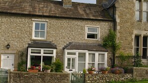 A stone house with a white door, two windows with shutters, and a small porch with potted plants. - Country Hideaways | Barton Cottage | West Burton (Leyburn)
