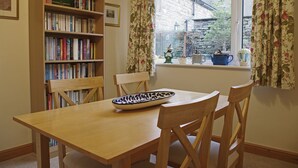 A dining area with a wooden table, chairs, a bookshelf, and a window with curtains. - Country Hideaways | Barton Cottage | West Burton (Leyburn)