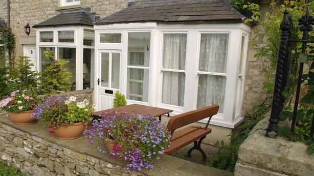 A stone building with a conservatory, a wooden bench, and potted plants. - Country Hideaways | Barton Cottage | West Burton (Leyburn)