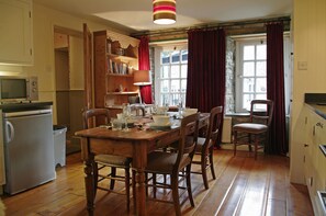 A wooden dining table set with plates, glasses, and cutlery. There is a refrigerator, a microwave, and a bookshelf in the room.