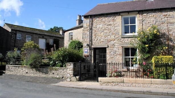 A stone building with a traditional design, featuring a small front porch and a black metal gate. - Country Hideaways | Jesmond Cottage | West Burton (Leyburn)