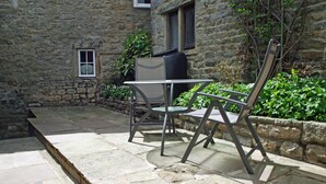 A stone building with a small window, a patio with stone flooring, and a metal chair and table. - Country Hideaways | Jesmond Cottage | West Burton (Leyburn)