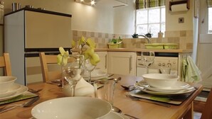 A kitchen with a dining table set for four, a refrigerator, a stove, and a window with curtains. - Country Hideaways | Jesmond Cottage | West Burton (Leyburn)