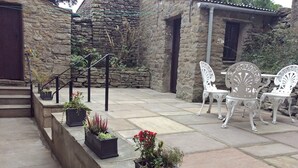 Un edificio de piedra con una escalera que da a un patio. Hay plantas en macetas y mobiliario exterior blanco en el patio.