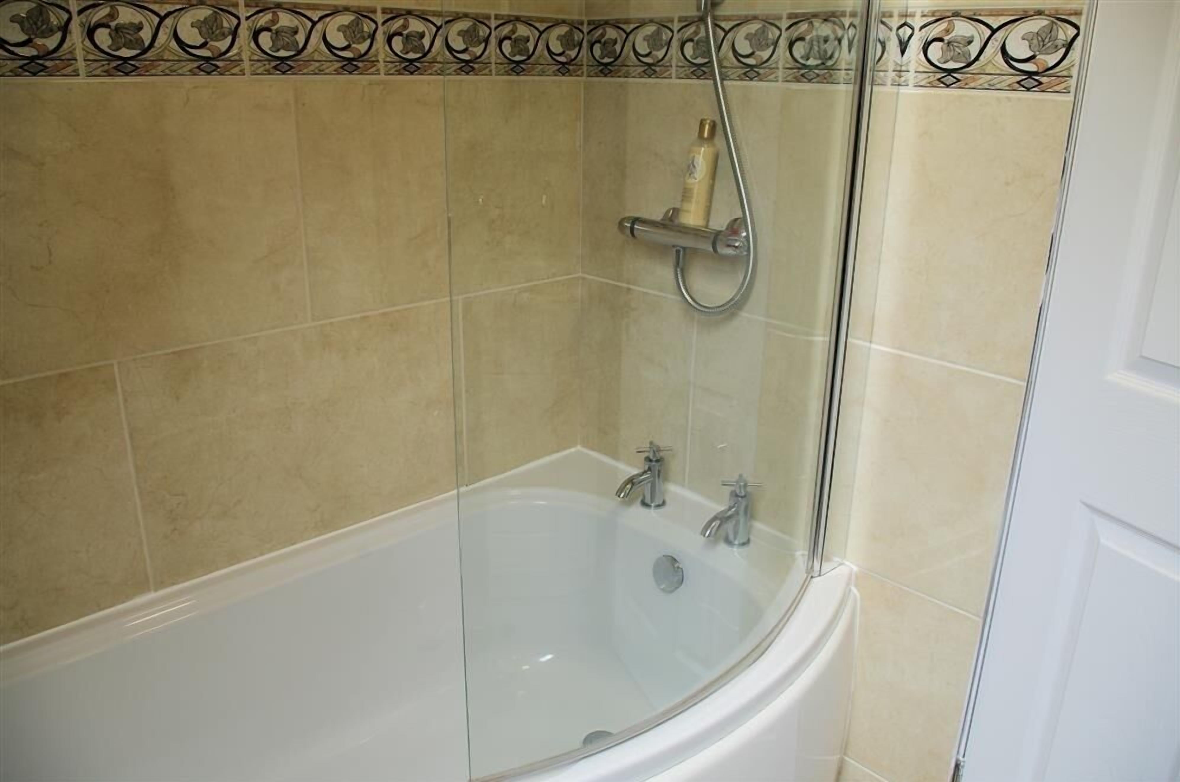 Een badkamer met een glazen badkuip, een douchekop en een decoratieve tegelrand.