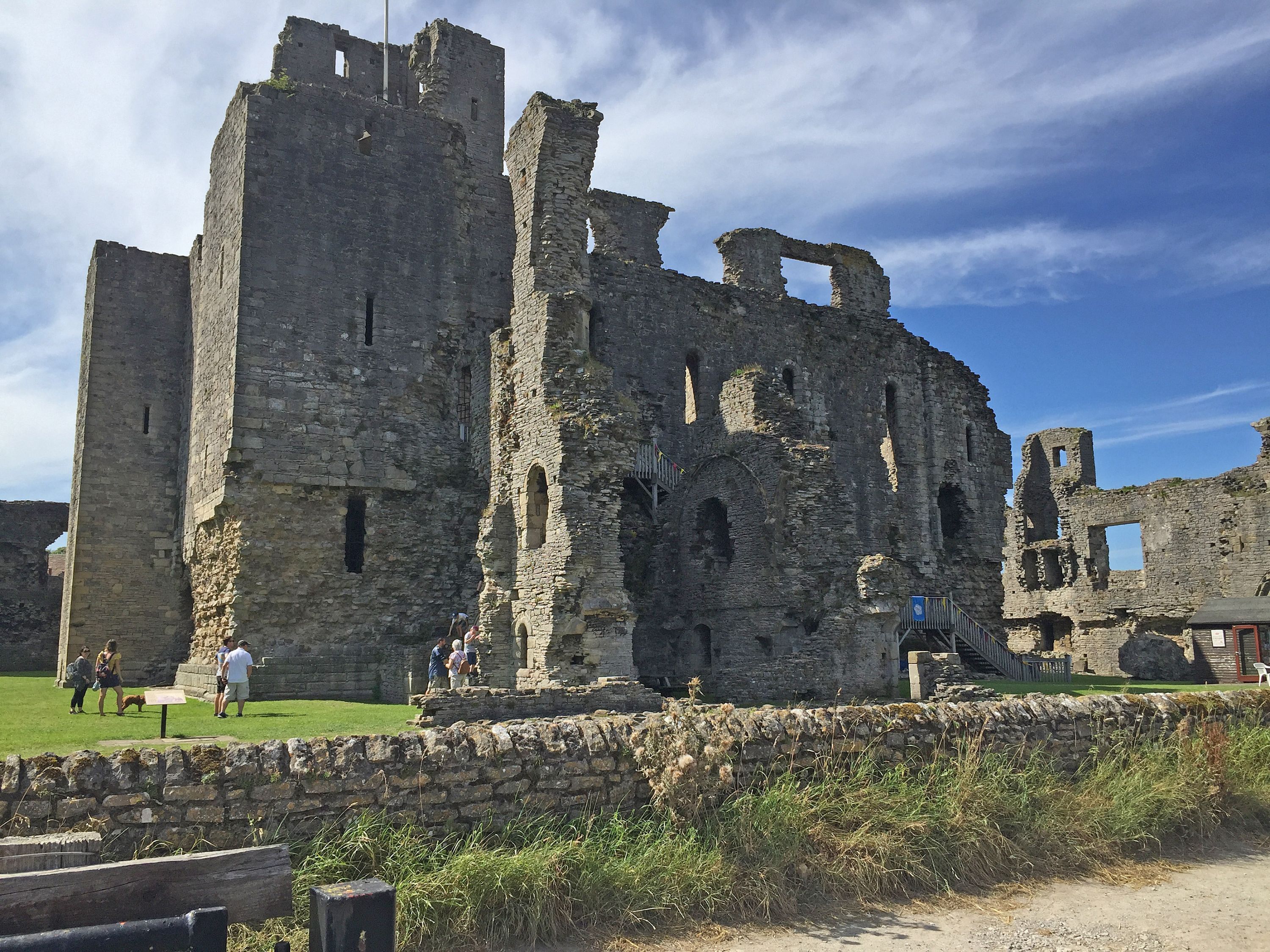 An ancient stone castle with multiple towers and arched windows, surrounded by a stone wall and a grassy area.
