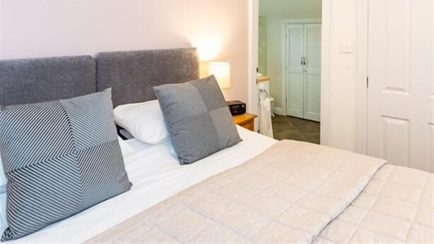 A neatly made bed with a white duvet and two pillows, a bedside table with a lamp, and a door leading to a bathroom.
