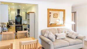 A living room with a beige sofa, wooden dining table, and chairs. A kitchen is visible in the background.