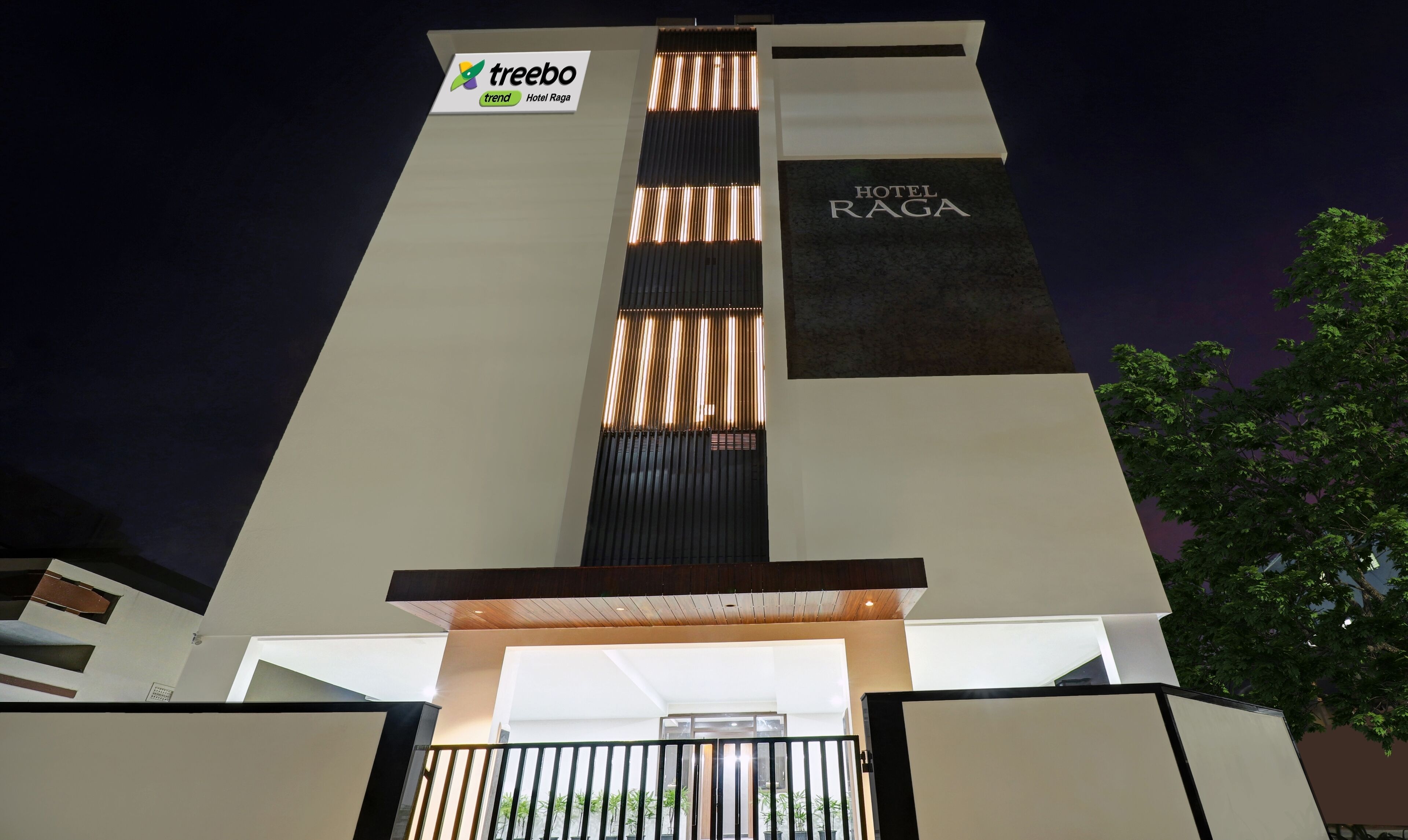 A modern hotel entrance with a vertical light feature, a sign for 'Treebo Trend Hotel Raga', and a 'HOTEL RAGA' sign.