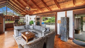A spacious living area with a wooden ceiling, a large gray sectional sofa, a wooden coffee table, wicker chairs, and a stone accent wall. - Hale Joli: Historic Hawaiian Home w/Ocean Views, Lanai & BBQ Area (Kailua-Kona)