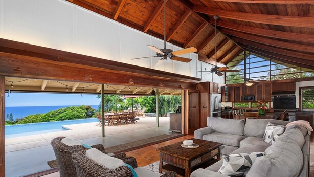 A spacious living area with a large gray sofa, wicker chairs, and a wooden coffee table. A ceiling fan and a view of a pool and lush greenery are visible. - Hale Joli: Historic Hawaiian Home w/Ocean Views, Lanai & BBQ Area (Kailua-Kona)