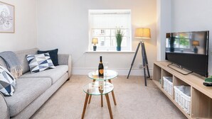 A modern living room with a grey sofa, a glass coffee table, a floor lamp, a TV on a wooden stand, and a window with blinds and a plant. - 1 bedroom accommodation in Helmsley (Helmsley)